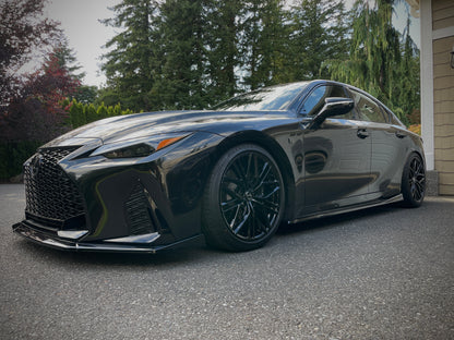 A glossy black lexus is500 nia splitter lip body kit luxury sedan parked on a driveway in front of trees and a house, shown from a three-quarter front angle.